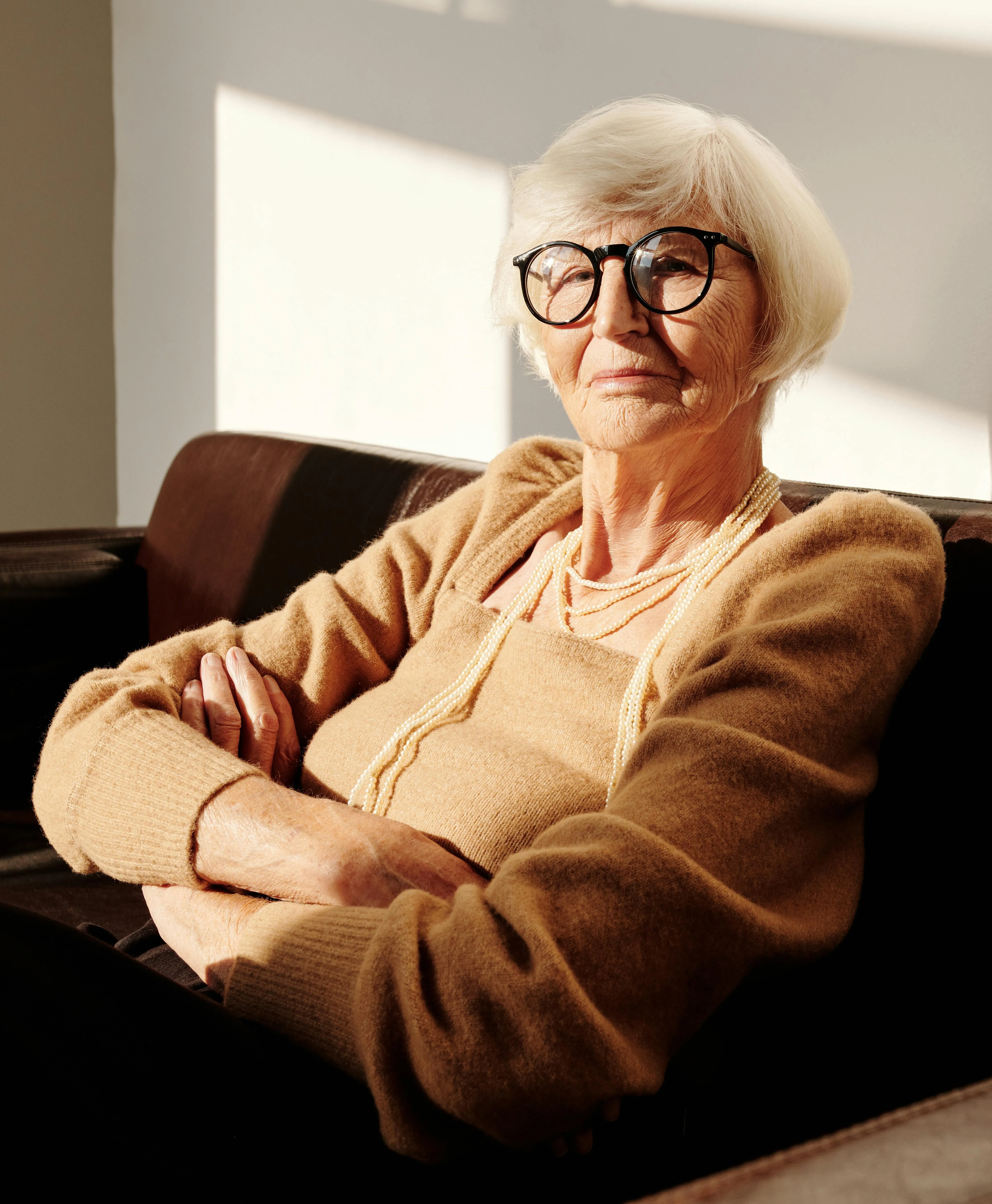 Пожилая женщина в очках сидит на диване, сложив руки